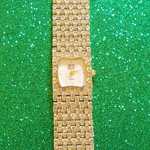May include: A gold-toned Nolan Miller quartz watch with a rectangular face and a band encrusted with small, sparkling stones. The watch face is white with gold hands and the brand name. The watch is displayed against a green, glittery background.