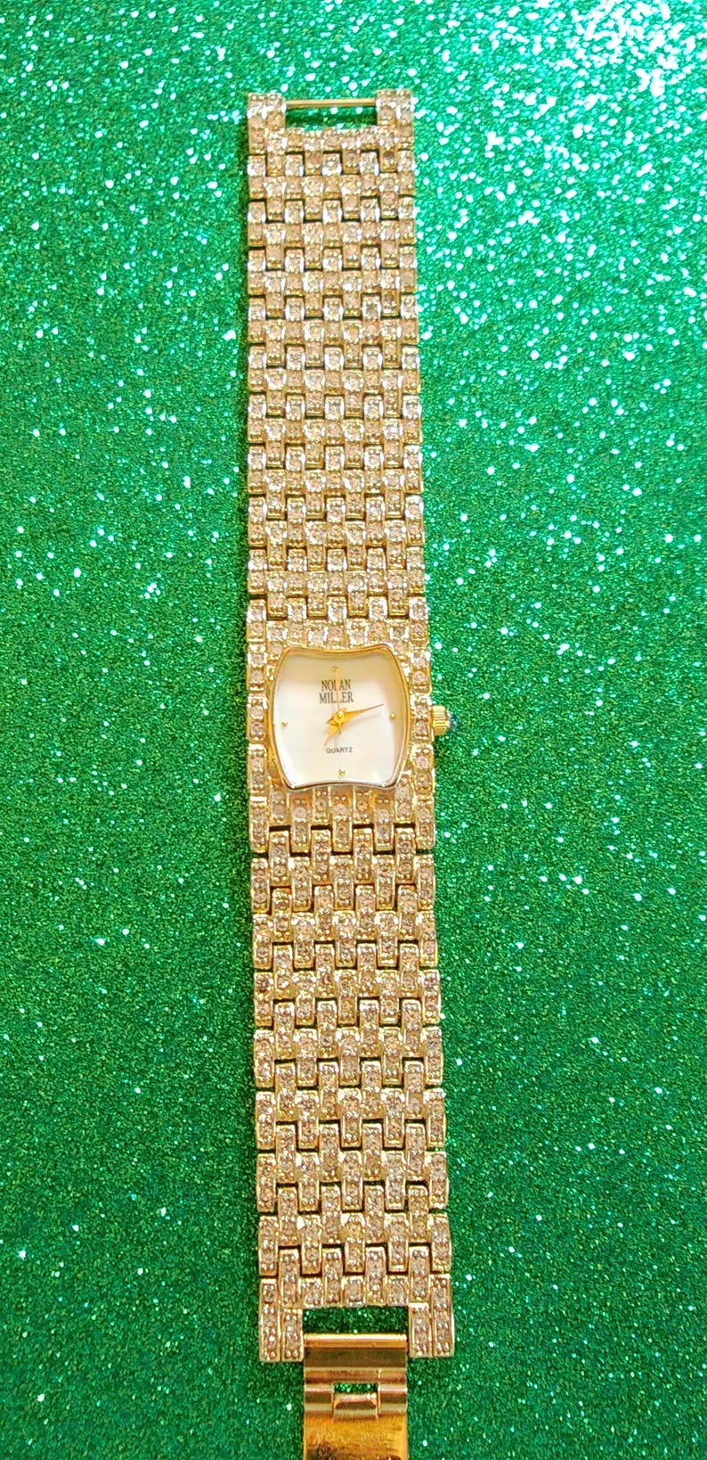 May include: A gold-toned Nolan Miller quartz watch with a rectangular face and a band encrusted with small, sparkling stones. The watch face is white with gold hands and the brand name. The watch is displayed against a green, glittery background.