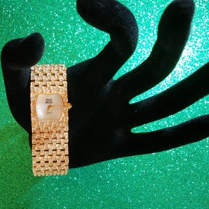 May include: A gold-toned wristwatch with a rectangular face and a band encrusted with small, sparkling stones. The watch face is white with gold hands and the brand name "Solan Miller". The watch is displayed on a black velvet hand against a green background.