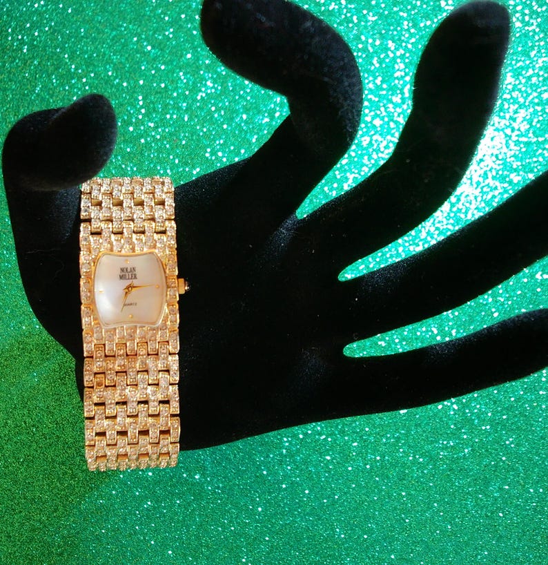 May include: A gold-toned wristwatch with a rectangular face and a band encrusted with small, sparkling stones. The watch face is white with gold hands and the brand name "Solan Miller". The watch is displayed on a black velvet hand against a green background.