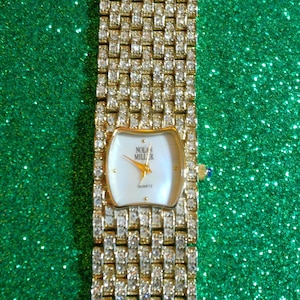 May include: A gold-toned quartz watch by Nolan Miller, featuring a rectangular face with a white dial and gold hands. The watch band is encrusted with sparkling crystals. The watch has a blue gem on the side.