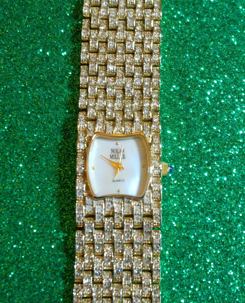 May include: A gold-toned quartz watch by Nolan Miller, featuring a rectangular face with a white dial and gold hands. The watch band is encrusted with sparkling crystals. The watch has a blue gem on the side.