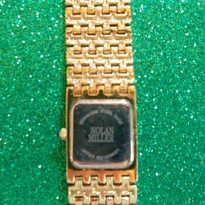 May include: Gold-toned Nolan Miller wristwatch with a rectangular face and a linked band. The watch face is black with white text. The band and watch case have a shiny finish. The watch is set against a sparkly green background.