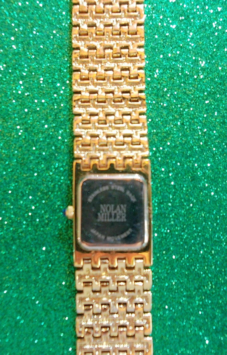 May include: Gold-toned Nolan Miller wristwatch with a rectangular face and a linked band. The watch face is black with white text. The band and watch case have a shiny finish. The watch is set against a sparkly green background.