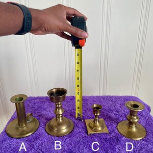 May include: Four brass candle holders of varying designs are displayed on a purple surface. A measuring tape shows the tallest candle holder is approximately 8 inches tall. The candle holders are labeled A, B, C, and D.
