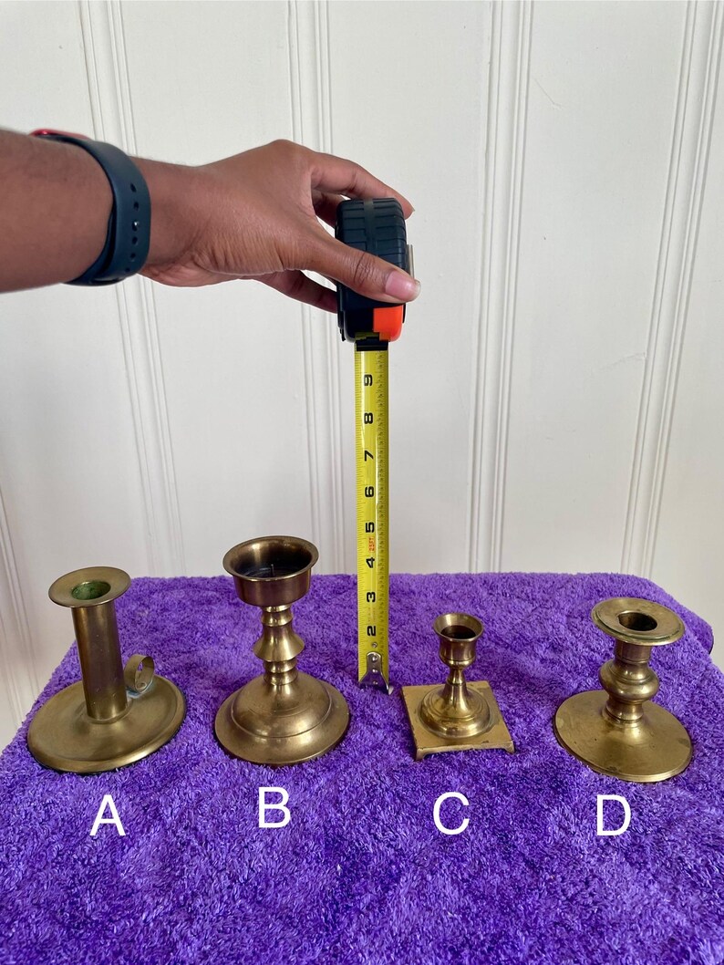 May include: Four brass candle holders of varying designs are displayed on a purple surface. A measuring tape shows the tallest candle holder is approximately 8 inches tall. The candle holders are labeled A, B, C, and D.