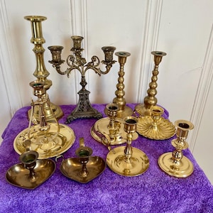 May include: A collection of brass candlesticks and candle holders in various styles. The assortment includes tall, ornate, and simple designs, with some featuring multiple candle holders. The pieces are arranged on a purple surface.