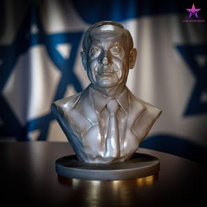 Puede incluir: Busto plateado de un hombre con traje y corbata, sobre una base redonda. La escultura está frente a una bandera israelí, con un fondo borroso. El busto tiene una superficie detallada y texturizada.