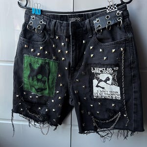 Könnte beinhalten: Schwarze Jeansshorts mit ausgefransten Kanten, verziert mit goldenen Nieten und Aufnähern. Ein Aufnäher zeigt eine grüne Grafik, der andere lautet: "I DIDN'T GO TO WORK TODAY... I DON'T THINK I'LL GO TOMORROW." Silberketten und Gürtelschlaufen mit Ringen.