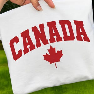 May include: White sweatshirt with the word "CANADA" in bold red letters and a red maple leaf emblem. The sweatshirt is held up against a green background.