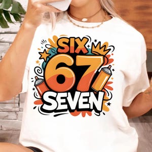 May include: White t-shirt with a colorful graphic design. The design features the words "SIX" and "SEVEN" in orange and white, with the number "67" in a large orange font. Includes two orange spray paint cans and a crown.