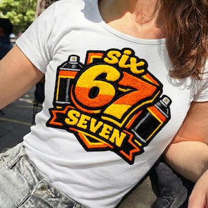 May include: White t-shirt featuring a colorful graphic design. The design includes the number "67" in orange and yellow, the word "SIX" above, and "SEVEN" below, with two spray paint cans. The design is outlined in black.