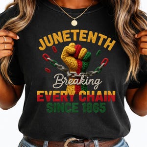 Op de afbeelding: Een zwart t-shirt met een afbeelding van een opgeheven vuist in de kleuren geel, rood en groen, die een zilveren ketting doorbreekt. De tekst luidt JUNETENTH, Breaking EVERY CHAIN SINCE 1865 in gele, witte, rode en groene letters.