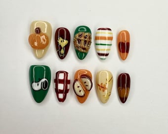 May include: A set of ten almond-shaped press-on nails with various autumn-themed designs. The designs include a 3D pumpkin, Snoopy, a gold wreath, stripes, and apple slices. Colors include orange, green, red, and yellow.