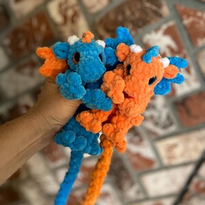 May include: Two plush, crocheted dragons in vibrant colors. One is blue with orange accents, the other is orange with blue accents. They have small, round eyes and long, curled tails. The dragons are soft and cuddly.