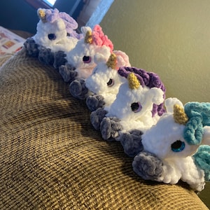 May include: A row of five handmade crochet unicorns in white, with gray bodies and manes and horns in various colors, including purple, pink, and blue. Each unicorn has large, round eyes.