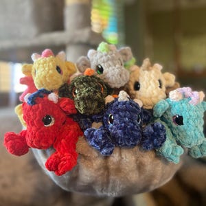 May include: A collection of handmade crochet dragons in various colors, including red, yellow, blue, and green. Each dragon has large, black eyes and small horns. The dragons are nestled together in a soft, gray basket.