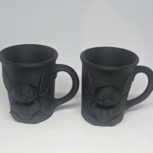 May include: Two black ceramic mugs with handles. Each mug features a raised relief of a cartoon character. The mugs are identical in shape and size, with a smooth, matte finish. The mugs are suitable for hot or cold beverages.