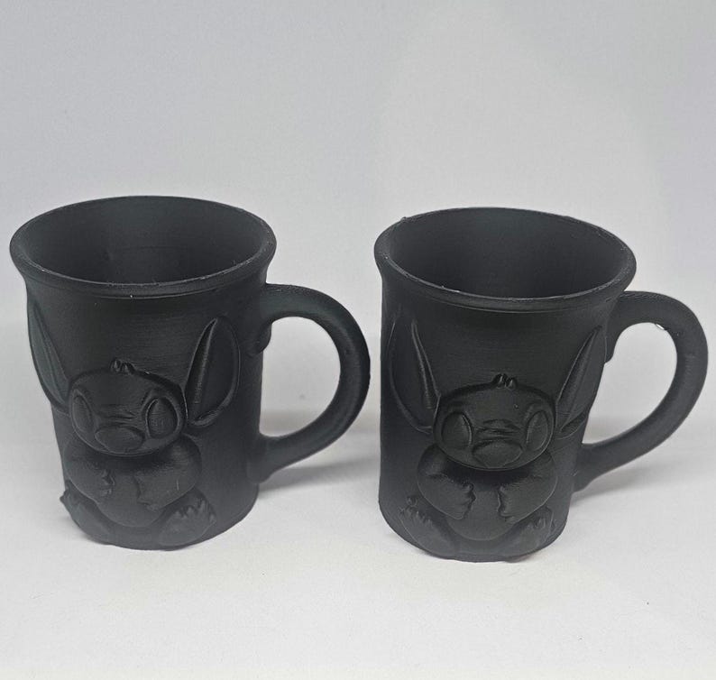 May include: Two black ceramic mugs with handles. Each mug features a raised relief of a cartoon character. The mugs are identical in shape and size, with a smooth, matte finish. The mugs are suitable for hot or cold beverages.
