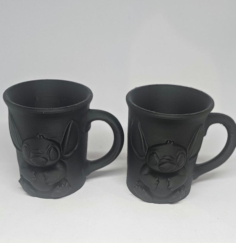 May include: Two black ceramic mugs featuring a raised design of a cartoon character. Each mug has a rounded handle and a cylindrical shape. The character design is on the front of each mug.