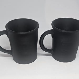 May include: Two black ceramic mugs with curved handles. The mugs have a slightly textured surface and a simple, minimalist design. The mugs are suitable for coffee, tea, or other beverages.