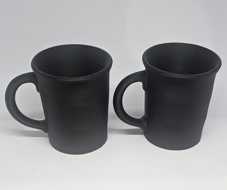 May include: Two black ceramic mugs with curved handles. The mugs have a slightly textured surface and a simple, minimalist design. The mugs are suitable for coffee, tea, or other beverages.