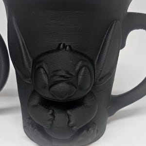 May include: Black mug featuring a 3D-printed design of a cartoon character. The mug has a handle and a textured surface. The character is in a sitting position with large ears and eyes.