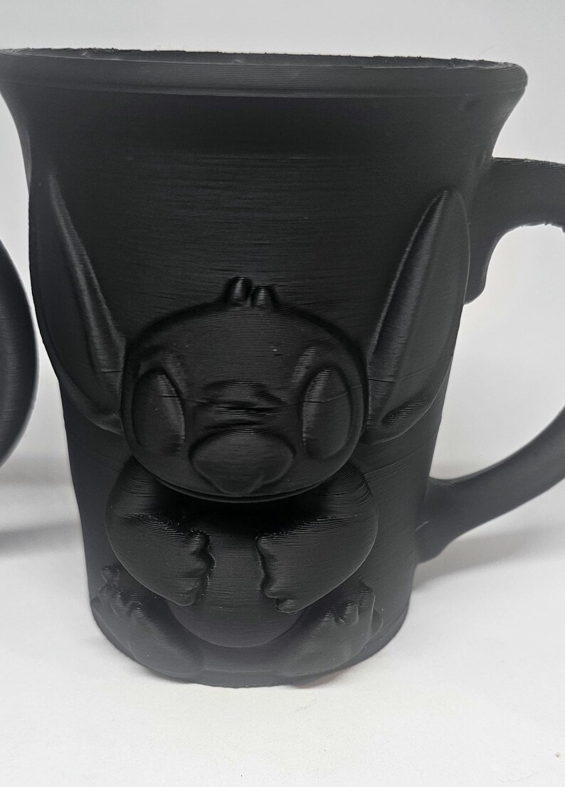 May include: Black mug featuring a 3D-printed design of a cartoon character. The mug has a handle and a textured surface. The character is in a sitting position with large ears and eyes.