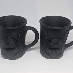 May include: Two black ceramic mugs with a raised design of a cartoon character. Each mug has a curved handle and a cylindrical shape. The character's face and body are visible on the front of the mugs.