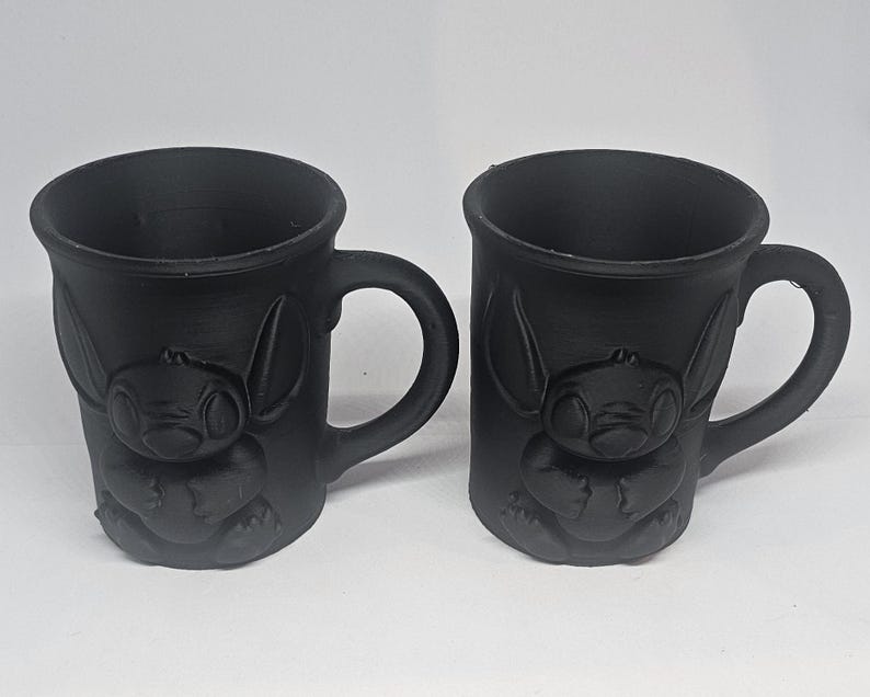 May include: Two black ceramic mugs with a raised design of a cartoon character. Each mug has a curved handle and a cylindrical shape. The character's face and body are visible on the front of the mugs.