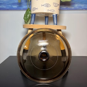 May include: A clear, brown glass lid with a metal handle. The lid is circular and sits on a dark surface. In the background, a white ceramic pot with blue legs and fish illustrations holds a plant.
