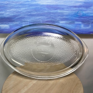 May include: Clear glass oval baking dish with a textured interior and a clear lid. The dish has a logo that reads "Made in Canada" and the brand name "Geward". The dish is sitting on a wooden surface.