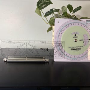 Puede incluir: Una regla de cálculo náutica de plástico transparente con una base de metal plateado y una regla de cálculo circular rosa y verde. La regla de cálculo tiene marcas de velocidad, tiempo y distancia. El texto "Weems & Plath" es visible.
