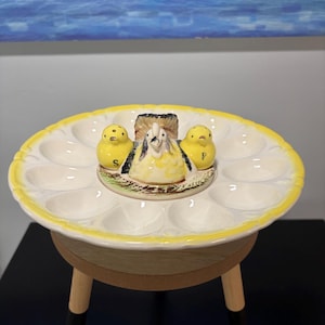 Op de afbeelding: Een keramische eierhouder met een gele rand en een middelpunt van een kip en kuiken. De eierhouder heeft 12 eiersleuven. Het middelpunt toont een kip en twee kuikens. De eierhouder staat op een kleine houten standaard.