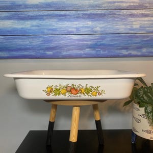 Puede incluir: Fuente rectangular blanca con borde negro y diseño colorido de frutas y verduras. La fuente se apoya en un taburete de madera con patas con punta negra. La palabra "Lechate" está impresa en la fuente.