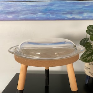May include: A clear glass lid sits atop a small, round, cork-topped table with three wooden legs. The legs are natural wood with black painted tips. The table is on a black surface, with a blue and white painting in the background.