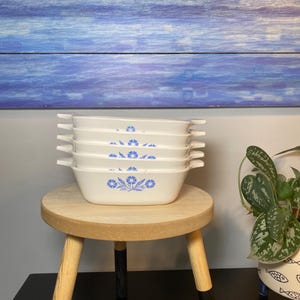 Puede incluir: Una pila de cinco fuentes rectangulares blancas con diseños florales azules. Cada fuente tiene dos asas y está apilada sobre un pequeño taburete redondo de madera. El fondo presenta un panel de madera pintado de azul y blanco.