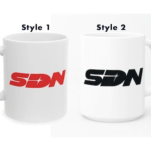 Puede incluir: Dos tazas de cerámica blanca con las letras "SDN" en una fuente estilizada. Una taza tiene letras rojas y la otra negras. Ambas tazas tienen un asa estándar y un acabado brillante. Los textos "Style 1" y "Style 2" están encima de las tazas.