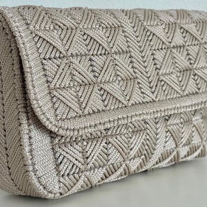 May include: A beige macrame clutch with a geometric pattern. The clutch has a flap closure and a woven texture. The bag is made of a soft, neutral-toned material, suitable for various occasions.