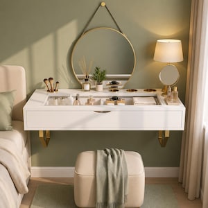May include: A white wall-mounted vanity table with a glass top, a single drawer, and gold-toned metal brackets. Above the table hangs a round gold-framed mirror. A beige upholstered ottoman with a draped green cloth sits below the vanity.