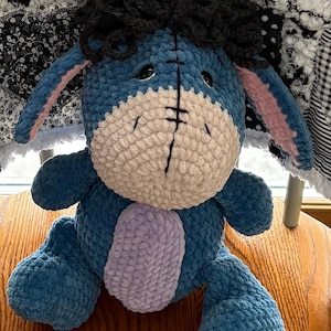 May include: A handmade blue plush donkey with a light pink inner ear, a light purple belly, and black yarn hair. The donkey has a cream-colored face and is sitting on a wooden surface. The plush toy is a character from a popular children's story.