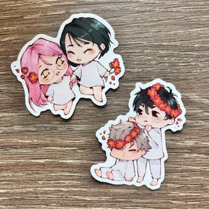 May include: Two sticker designs featuring chibi-style characters. One sticker shows a pair of characters holding hands, one with pink hair and glasses, the other with black hair. The second sticker shows two characters, one with gray hair and one with black hair, wearing flower crowns.