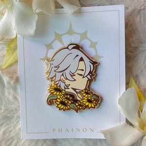 May include: A gold-colored enamel pin featuring a character with white hair and a sunflower collar, set against a white card with a gold border and a decorative pattern. The word "PHAINON" is printed at the bottom.