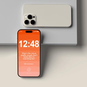 Peut inclure: Deux smartphones, l'un avec un écran orange affichant l'heure 12h48 et du texte sur la conscience de soi. L'autre téléphone est de couleur claire. Les téléphones sont sur une surface blanche.