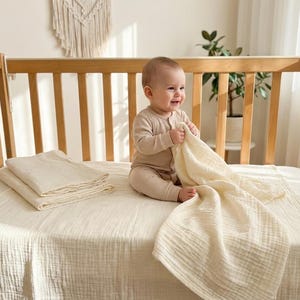 May include: A baby sits on a cot mattress, holding a cream-coloured muslin blanket. A stack of folded muslin blankets sits to the left. The cot is made of light-coloured wood.