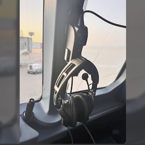 May include: A black aviation headset with a microphone hangs inside an aircraft cockpit. The headset is suspended from a black mount. The background shows a view of an airport. The image is taken from inside the cockpit.