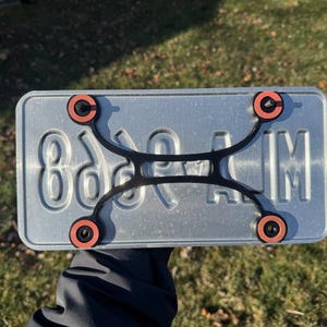 Pode incluir: Uma placa de matrícula prateada com letras e números em relevo, segurada contra um fundo de relva verde. Um suporte preto com quatro fixadores com anéis laranja fixa a placa. A superfície da placa é ligeiramente refletora.