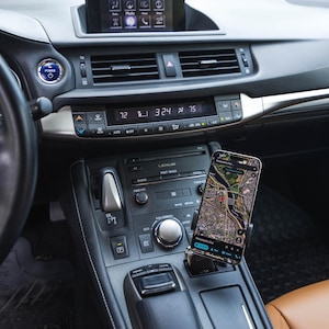 May include: A smartphone showing a map app is mounted in a car's console. The car's dashboard has a digital display, climate controls, and a radio. The interior is black and brown.