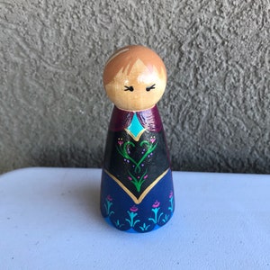 Peut inclure: Poup&eacute;e en bois peinte avec une t&ecirc;te marron clair et des cheveux marron clair. Les v&ecirc;tements de la poup&eacute;e sont peints avec une jupe bleu fonc&eacute;, un corsage noir et une cape violette. Le corsage a une bordure dor&eacute;e et un accent turquoise.