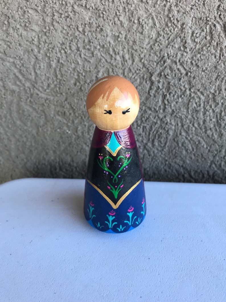 Peut inclure: Poup&eacute;e en bois peinte avec une t&ecirc;te marron clair et des cheveux marron clair. Les v&ecirc;tements de la poup&eacute;e sont peints avec une jupe bleu fonc&eacute;, un corsage noir et une cape violette. Le corsage a une bordure dor&eacute;e et un accent turquoise.